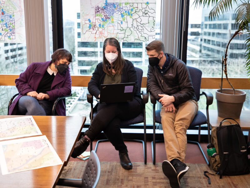 Three people wearing masks look at a laptop. Maps hang on the windows behind them.