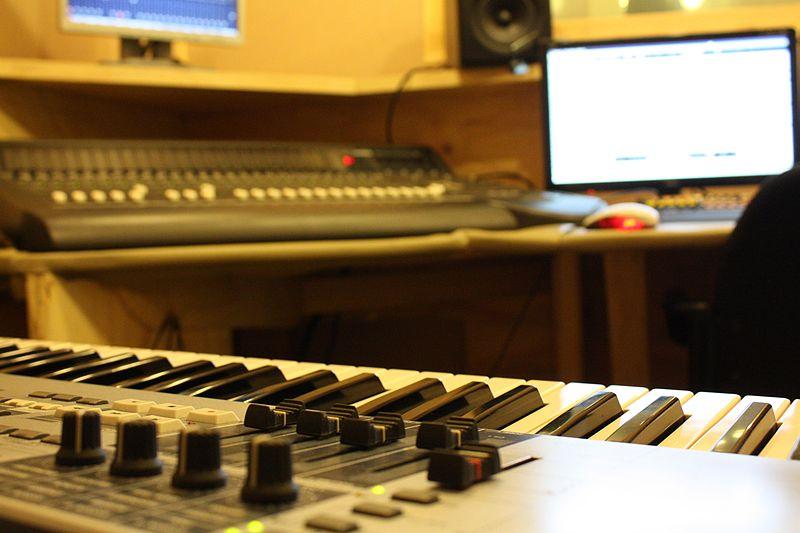 Recording studio with keyboard in foreground and computer and soundboard in background.