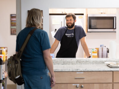 A person inside a kitchen wearing an apron talks through a window to another person.