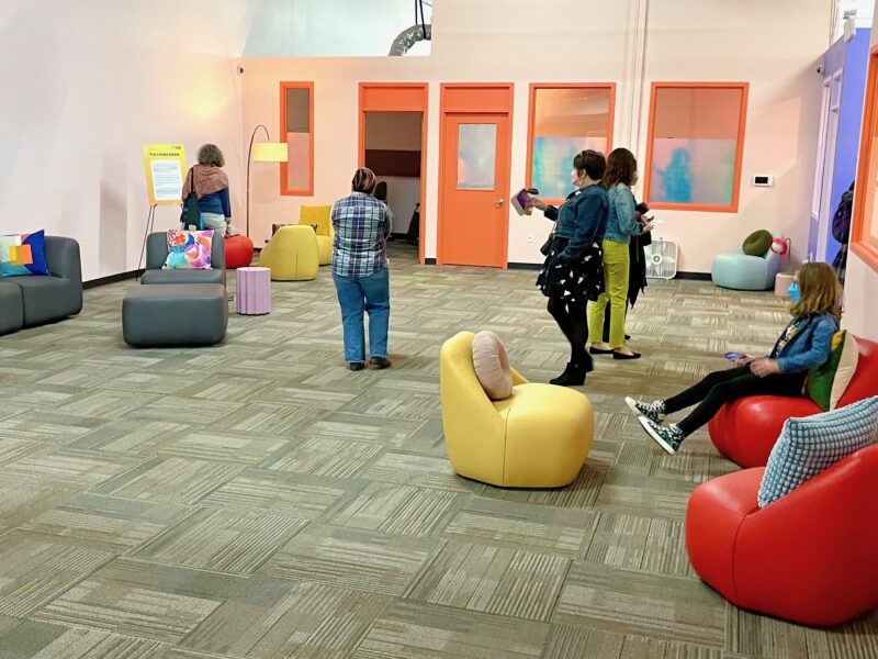 A large room with colorful furniture and art. People walking around.